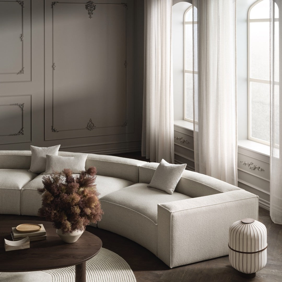 Curved beige sofa with light gray pillows, set against soft cream walls and sheer curtains, evoking modern elegance.