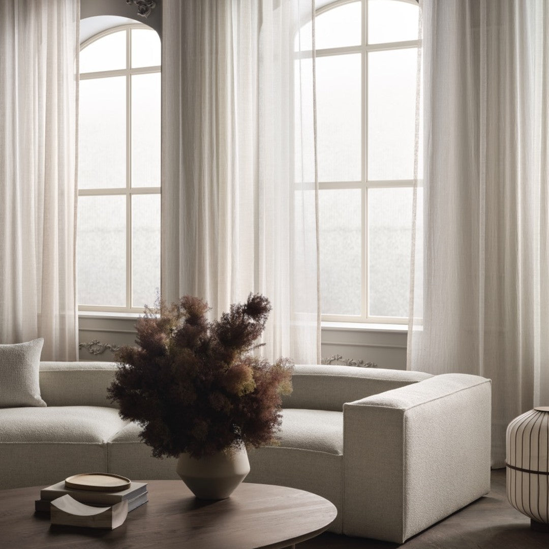 Light beige curtains and textured gray sofa with brown floral arrangement on a wooden table. Soft and neutral tones.