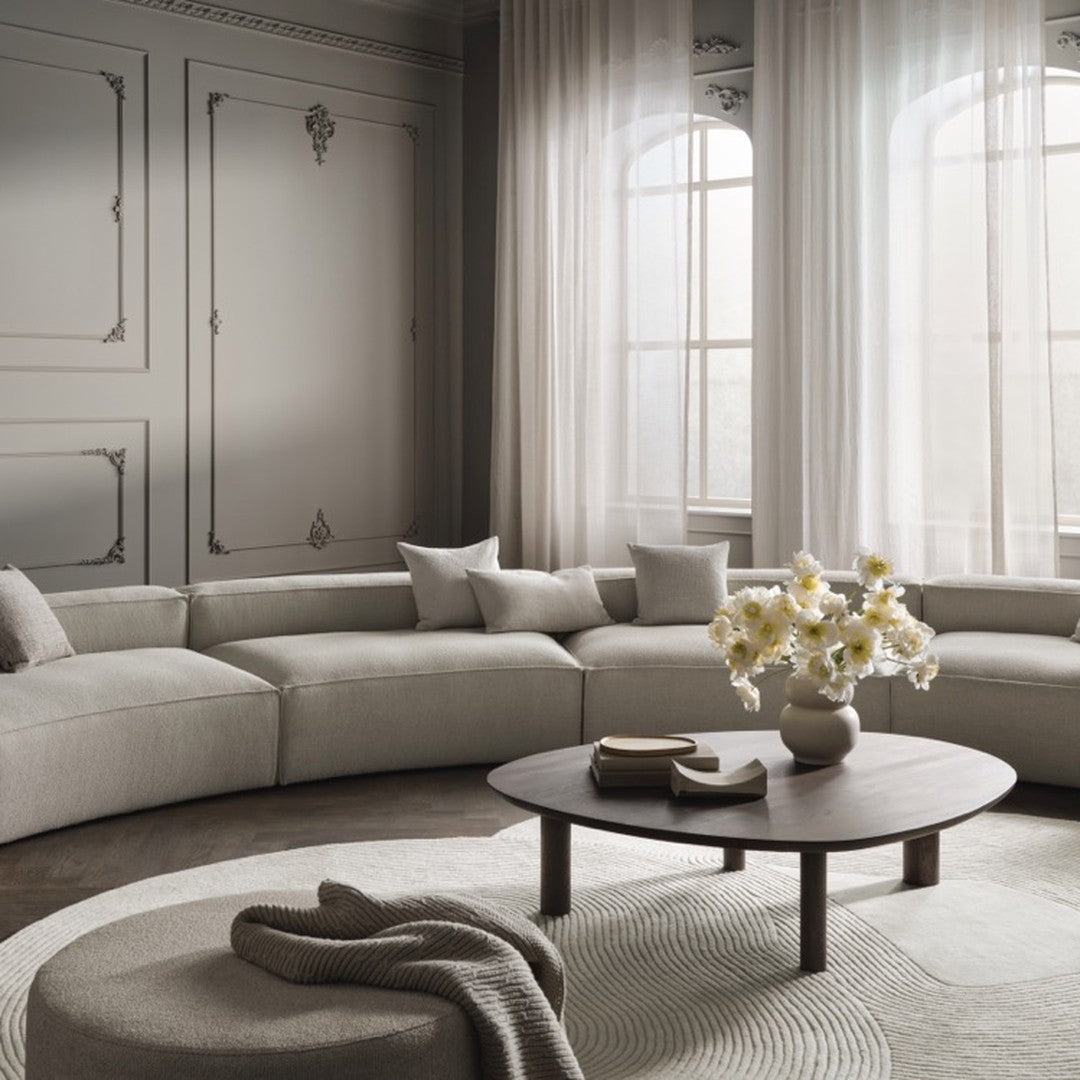 "Neutral beige sofa with gray walls and soft white curtains, accented by a round wooden table and patterned rug."