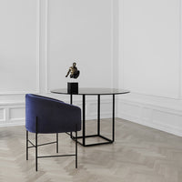 Modern round black table with a smooth surface, complemented by a deep blue upholstered chair, set on light wood herringbone floor.