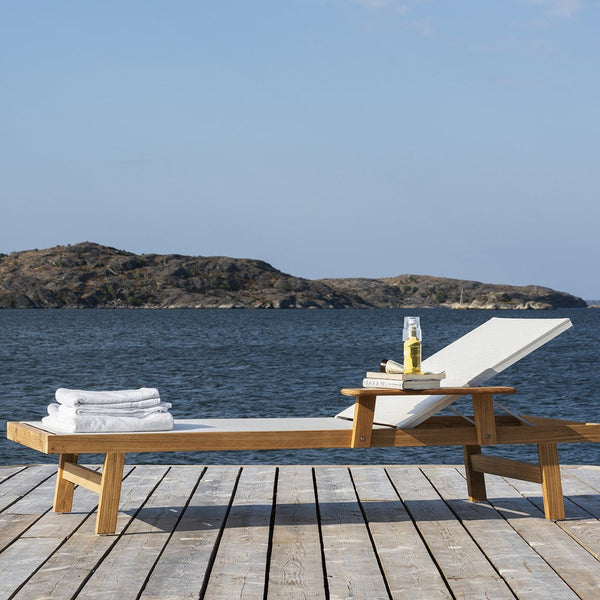 "White chaise lounge with natural wood frame, set against a blue water backdrop under a clear sky."