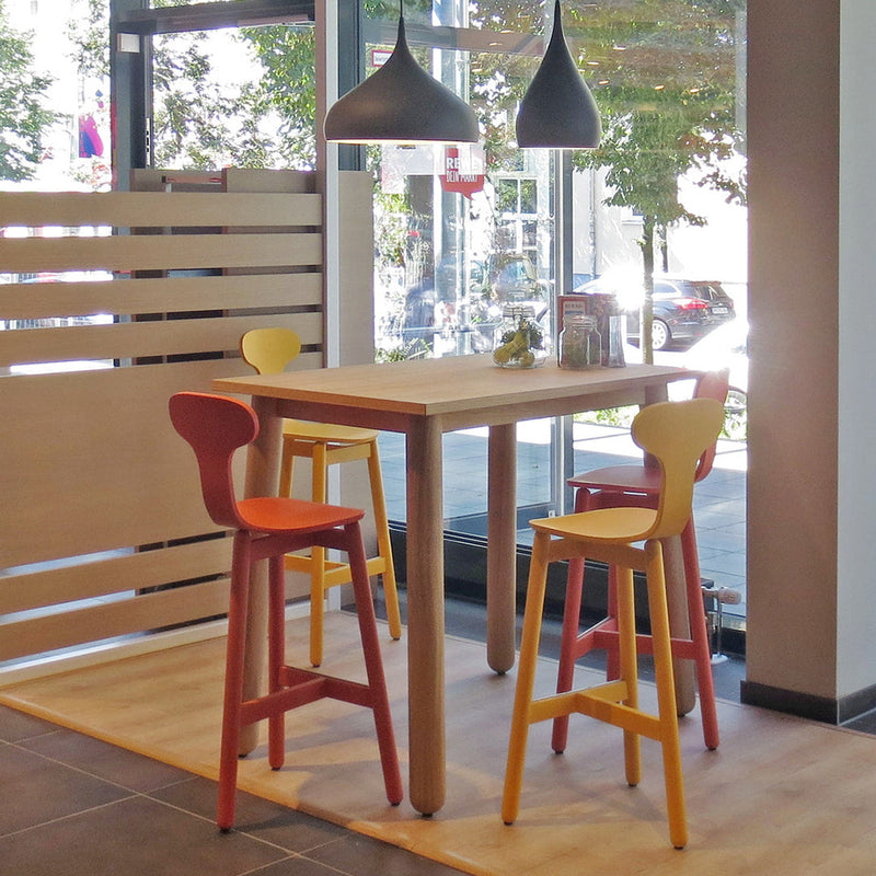 Colorful stools in red, yellow, and orange around a light wooden table in a modern cafe setting.