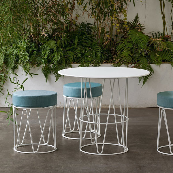 Modern white round table with geometric wire base and light blue upholstered stools among lush greenery.