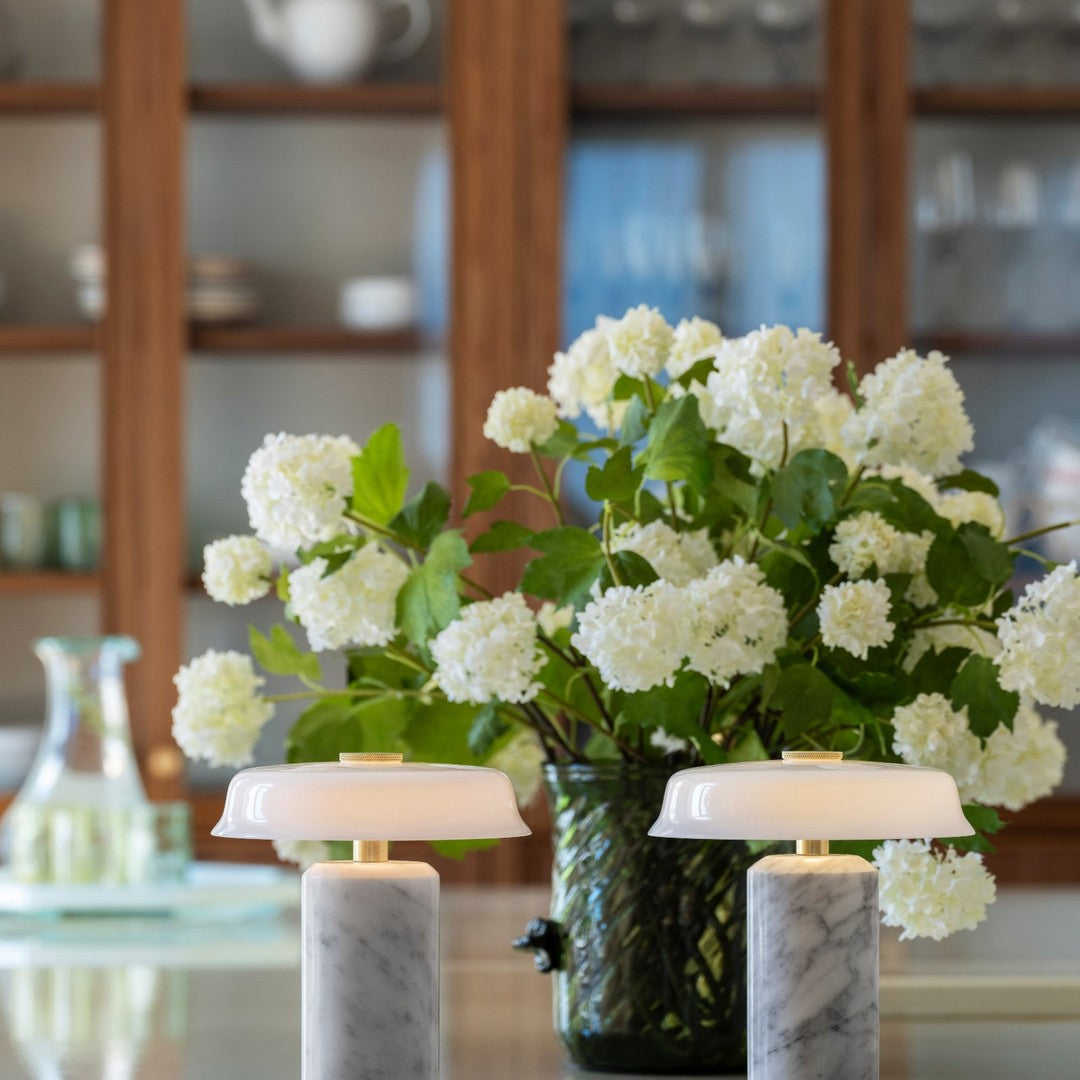 White floral arrangement in a dark green vase with two marble and white lamps on a glass table.