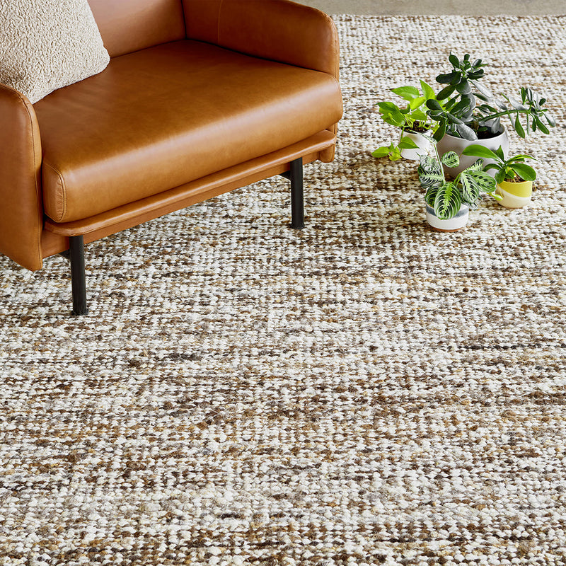 Brown and cream textured rug with subtle patterns, complementing a stylish brown leather chair and green plants.