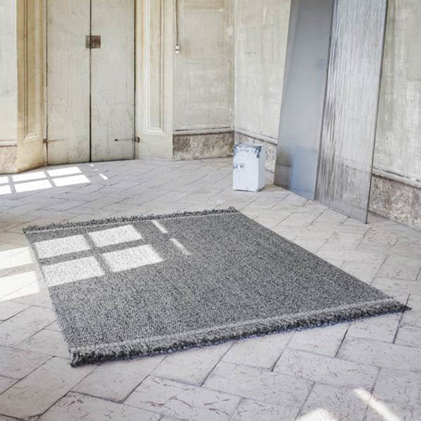 Gray textured rug with fringed edges, set against a light stone floor and minimalist background.