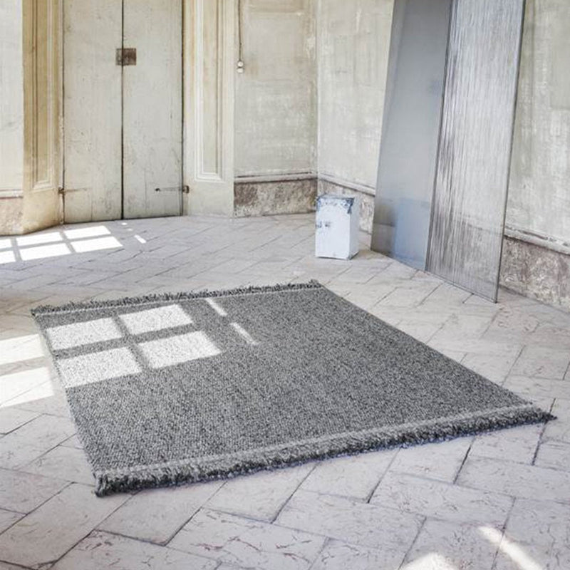 Gray textured rug with fringed edges, set against a light stone floor and minimalist background.