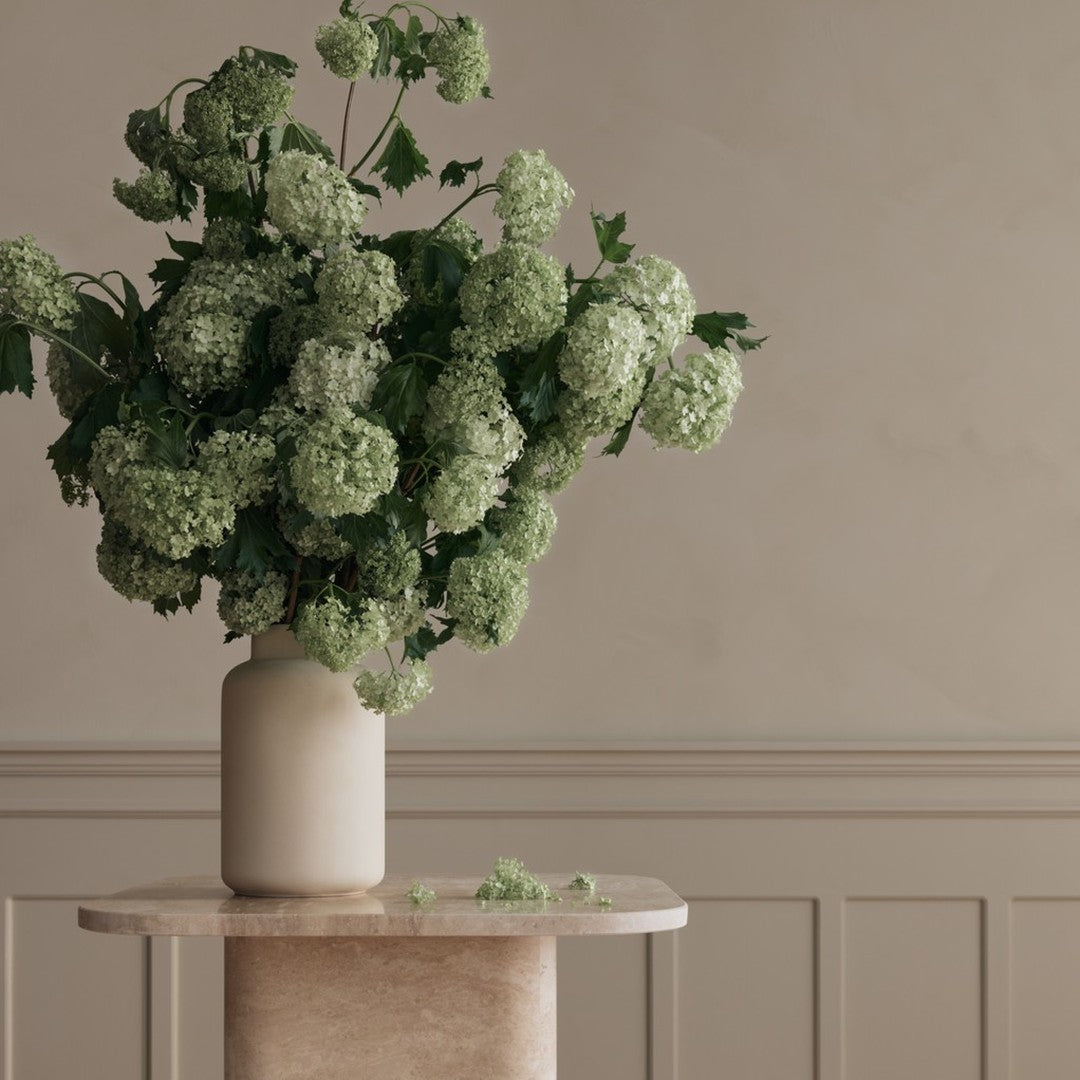 Bouquet of green flowers in a cream vase atop a light marble pedestal, set against a neutral background.