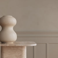 Beige sculptural vase on a light-colored marble pedestal against a neutral, textured wall. Elegant and modern design.