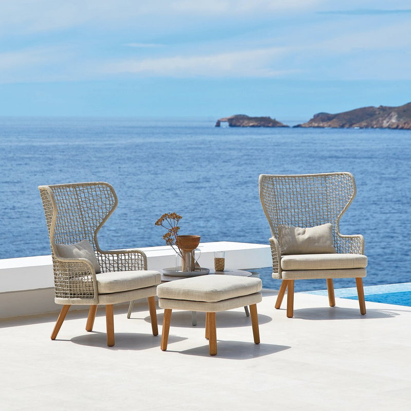 "Light beige woven chairs with cream cushions and natural wood legs, set against a serene blue sea backdrop."