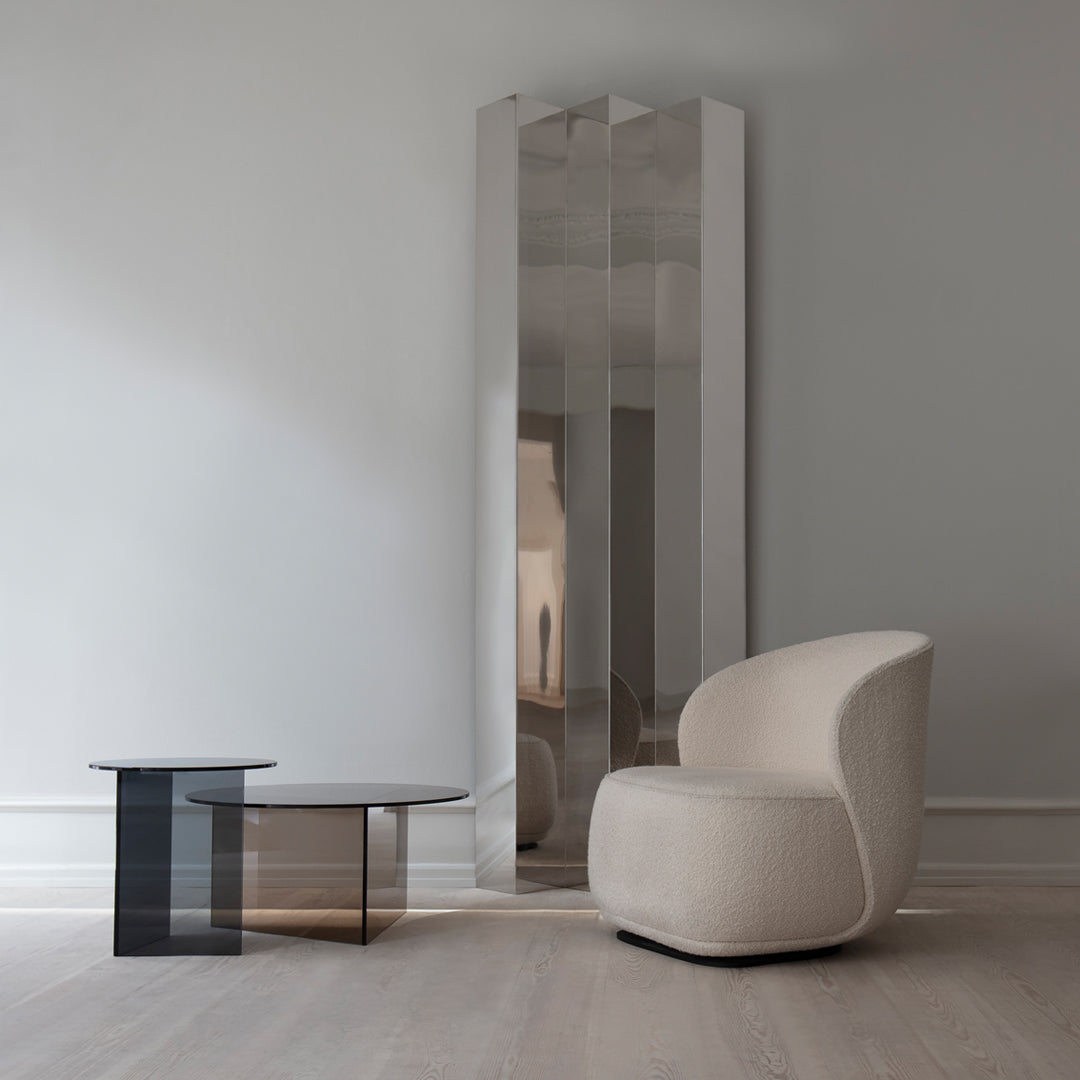 Modern beige chair and sleek black glass table beside a tall, reflective silver mirror against a light gray wall.