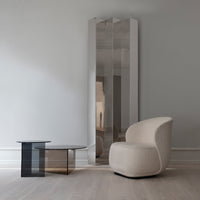 Modern beige chair and sleek black glass table beside a tall, reflective silver mirror against a light gray wall.