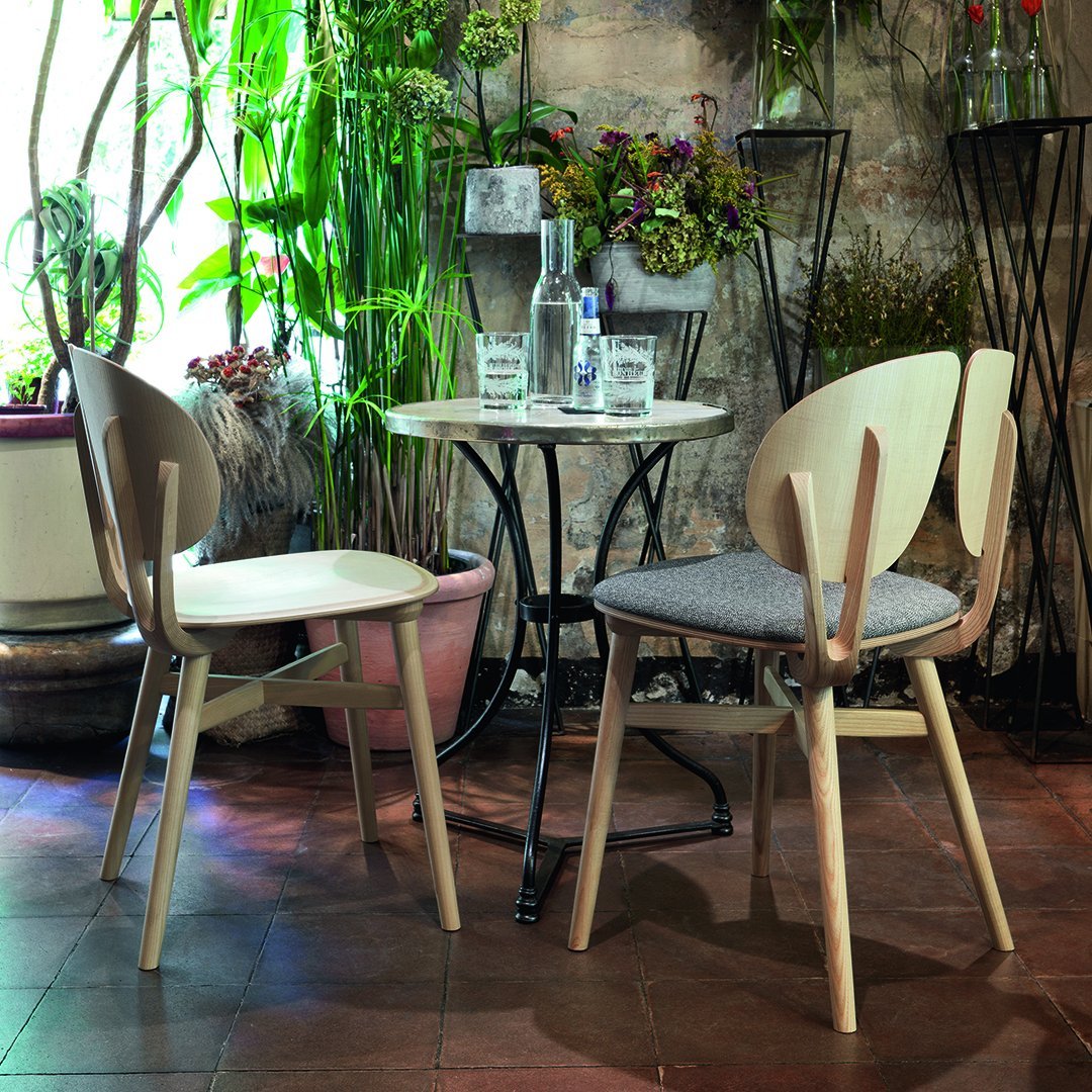 "Light wood chairs with gray upholstery around a black metal table, set in a lush green indoor garden."