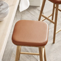 Tan padded seat with a smooth finish, complemented by light wooden legs on a textured floor.