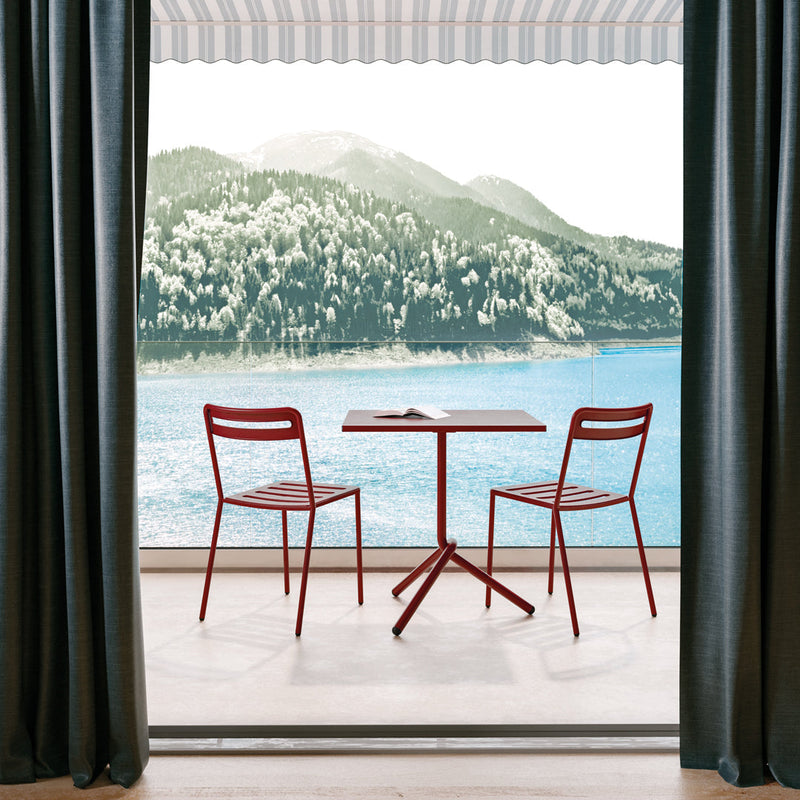 Red chairs and table in a serene outdoor setting, framed by dark curtains and striped awning.