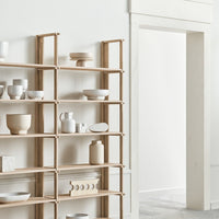 Light wood shelving unit displaying various white ceramic dishes and decor, against a neutral wall backdrop.