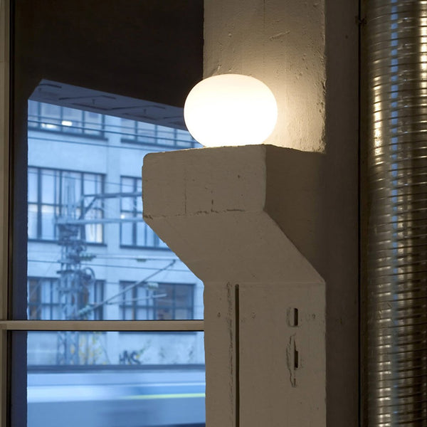 Minimalist white lamp with a round shade, set against a muted gray wall and urban window backdrop. 