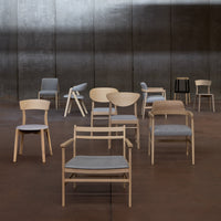 Collection of wooden chairs in light tones with gray cushions; varying designs and patterns on each chair.