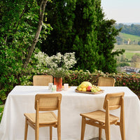"White tablecloth with natural wood chairs, set against lush greenery and a colorful fruit centerpiece."