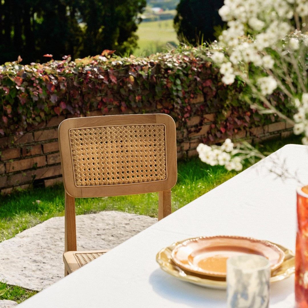 Natural wood chair with woven pattern, vibrant orange and white plates, surrounded by greenery and floral accents.