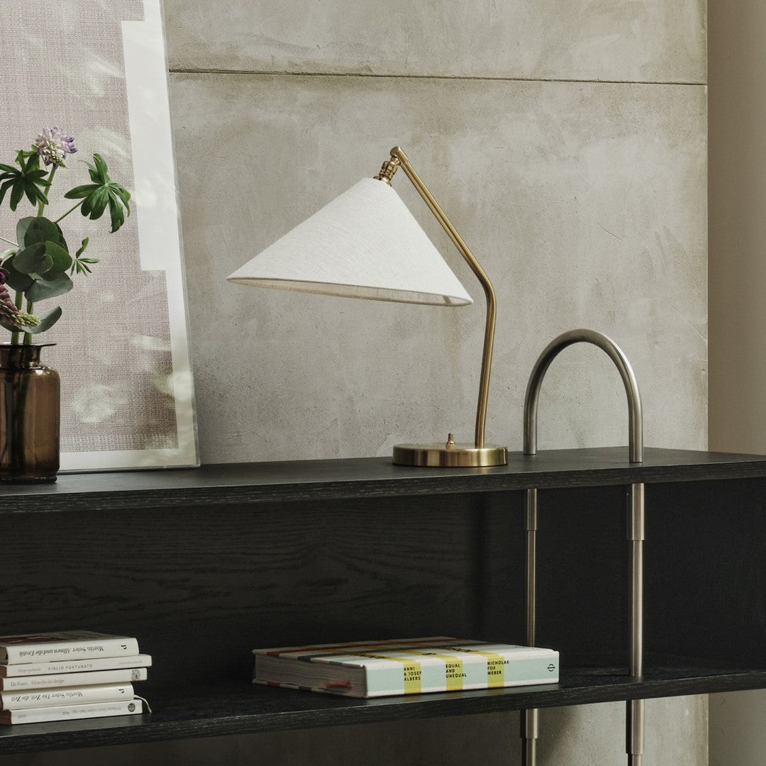 White conical lamp shade with brass accents, set against a gray concrete wall and dark wood shelf. Elegant modern design.