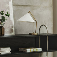 White conical lamp shade with brass accents, set against a gray concrete wall and dark wood shelf. Elegant modern design.