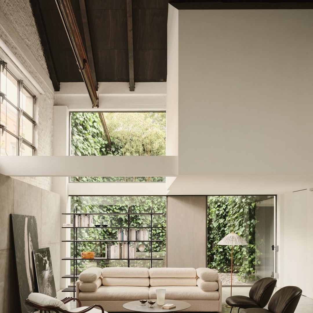 Minimalist living room with neutral beige sofa, dark wood accents, and lush green plants framing large windows. 