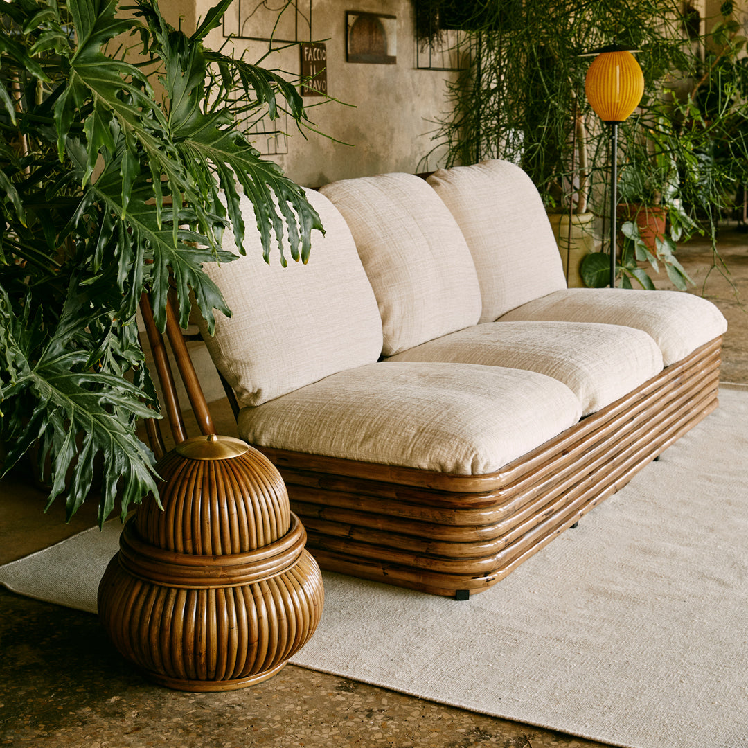 Beige and cream three-seat sofa with a textured fabric, paired with a brown, ribbed accent table, surrounded by greenery.