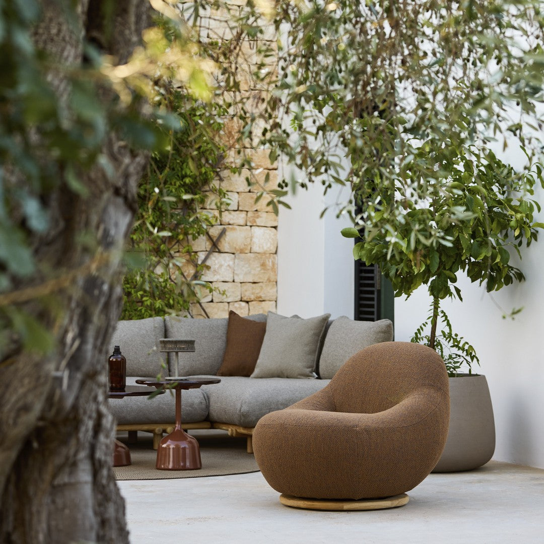 Brown organic-shaped chair with textured fabric, surrounded by gray cushions and greenery in a modern outdoor setting.