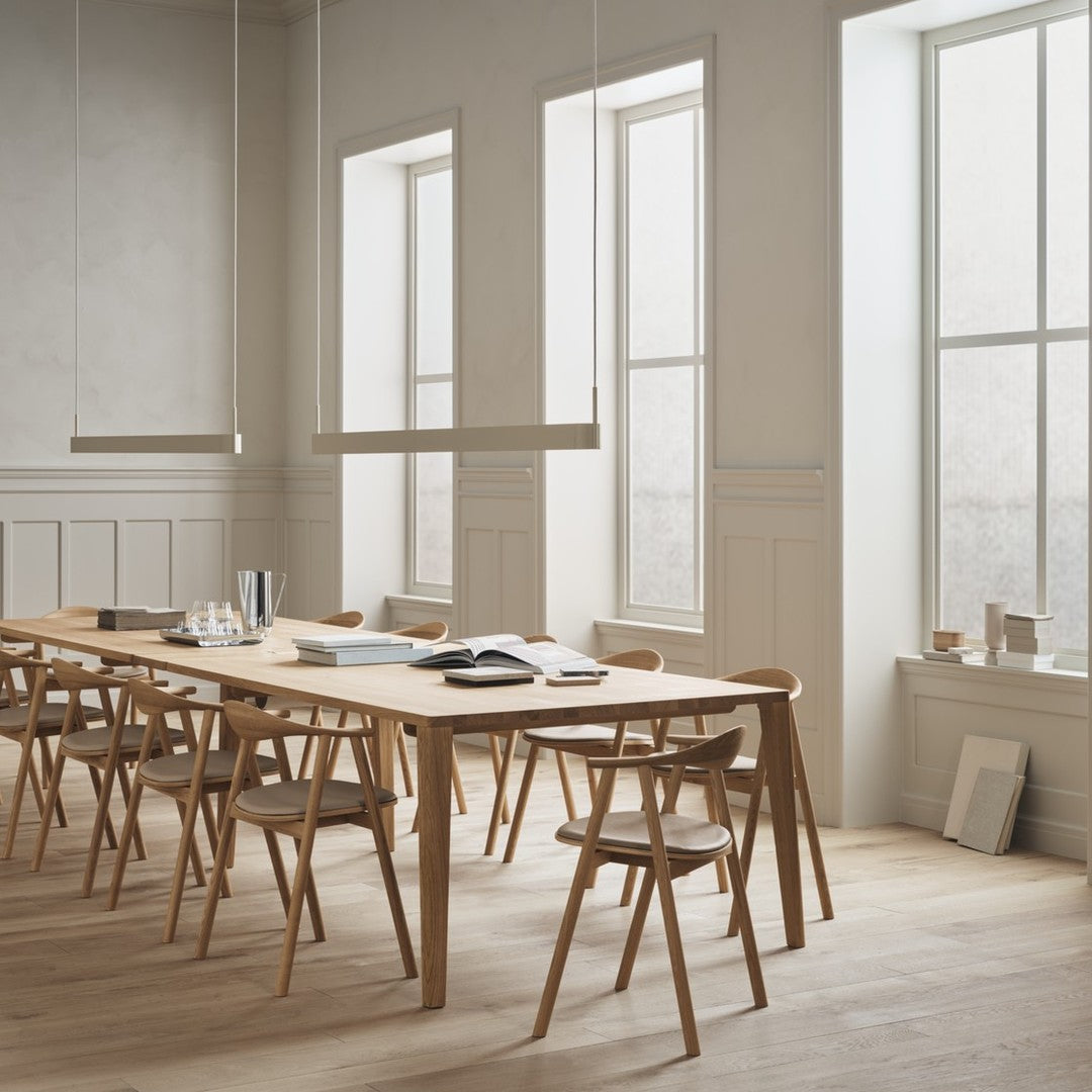 Light wood dining table and chairs in a minimalist room with soft, neutral tones and natural light from large windows.