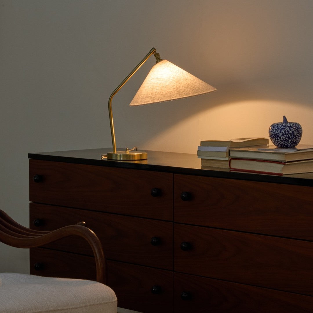 Brass desk lamp with a linen shade, set against a dark wood dresser. Subtle warm tones and a textured blue decorative piece.