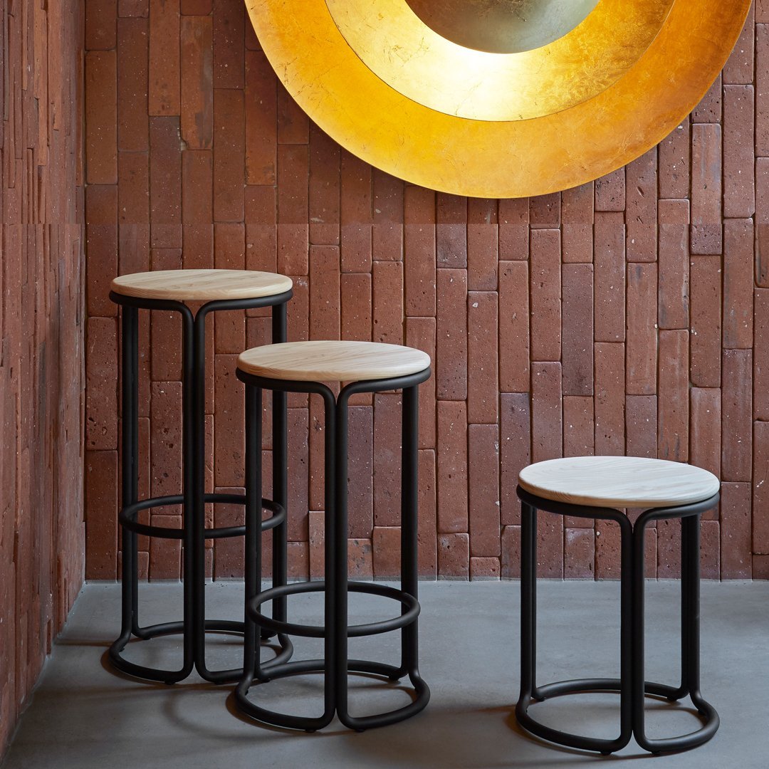Set of three modern bar stools featuring black metal frames and light beige cushions against a textured brick wall.