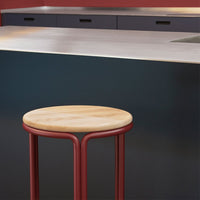 "Modern bar stool with a natural wood seat and sleek red metal legs, set against a darkly colored kitchen backdrop."