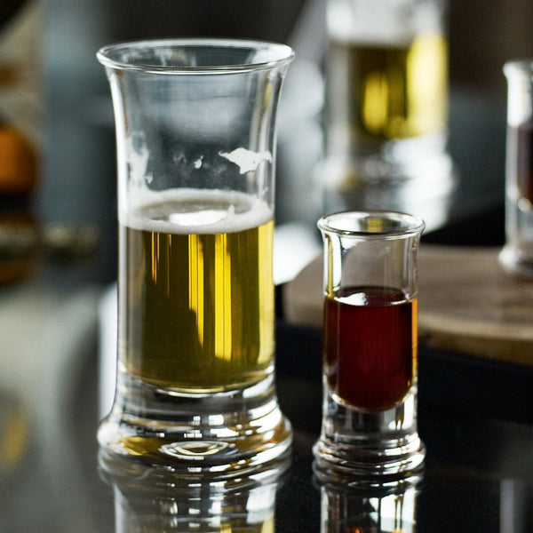 "Clear glass shot and beer glasses showcasing rich amber and golden hues, set against a sleek black surface."