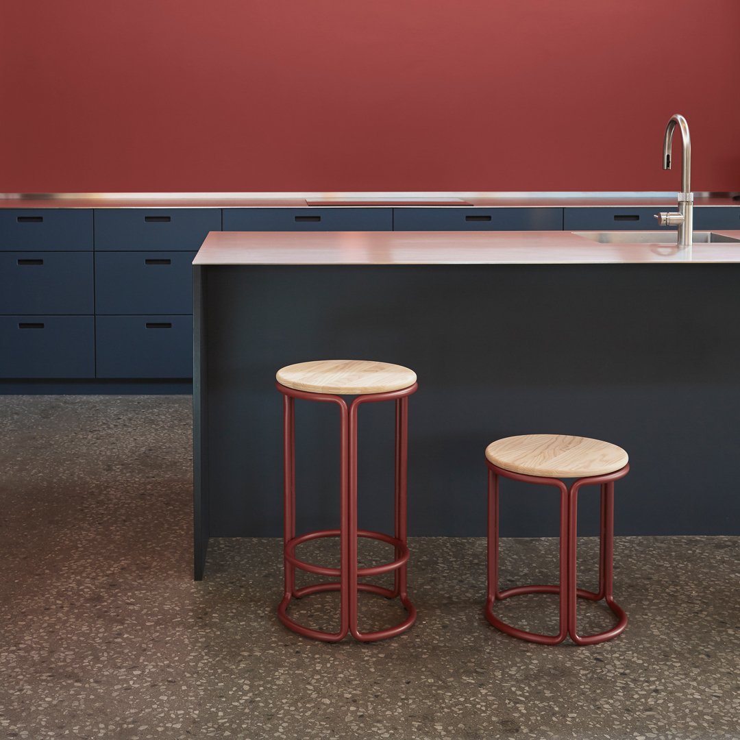 Two modern stools with natural wood seats and rust-colored metal frames, set against a deep red wall and navy cabinets.
