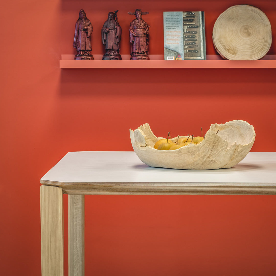 White tabletop with natural wood grain, vibrant orange wall, decorative wooden bowl with yellow fruits.