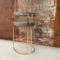 "Elegant gold metal bar stool with a sleek gray seat, showcased against a textured rust background."