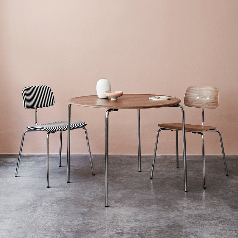Round wooden table with light brown finish, accompanied by gray and light brown striped chairs against a soft pink backdrop.