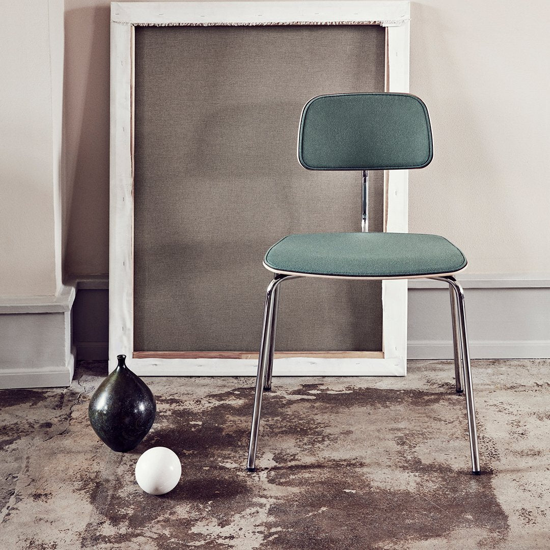 Modern teal chair with a sleek chrome frame, against a neutral backdrop and textured floor. Minimalist design elements.