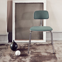 Modern teal chair with a sleek chrome frame, against a neutral backdrop and textured floor. Minimalist design elements.