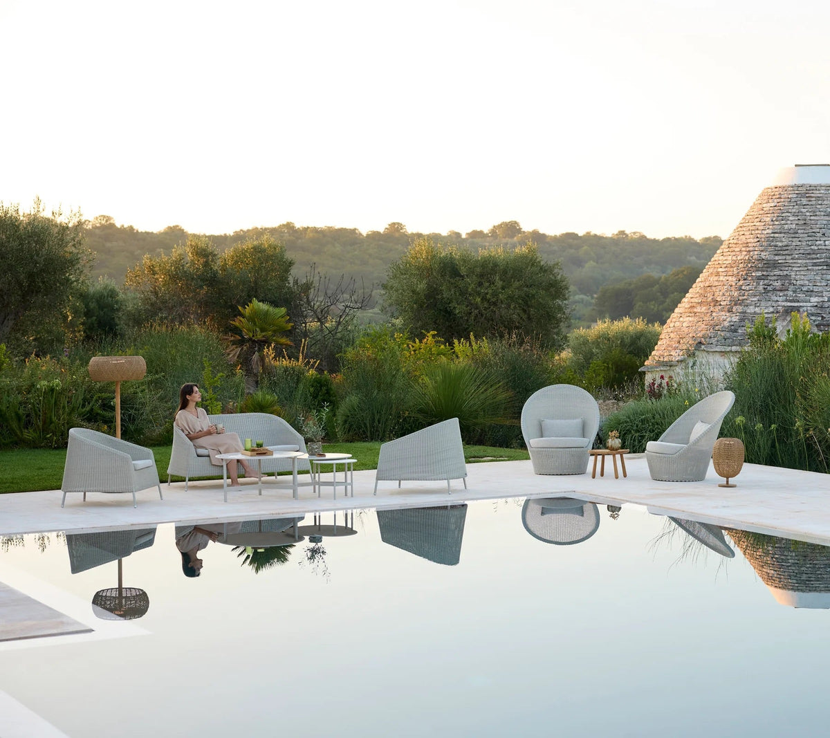 Elegant gray outdoor furniture with soft curves, set against lush green landscaping and serene water reflections.