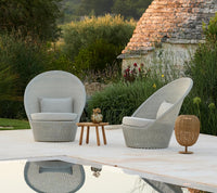 Light gray woven chairs with rounded backs, paired with a wooden side table, set against lush greenery.