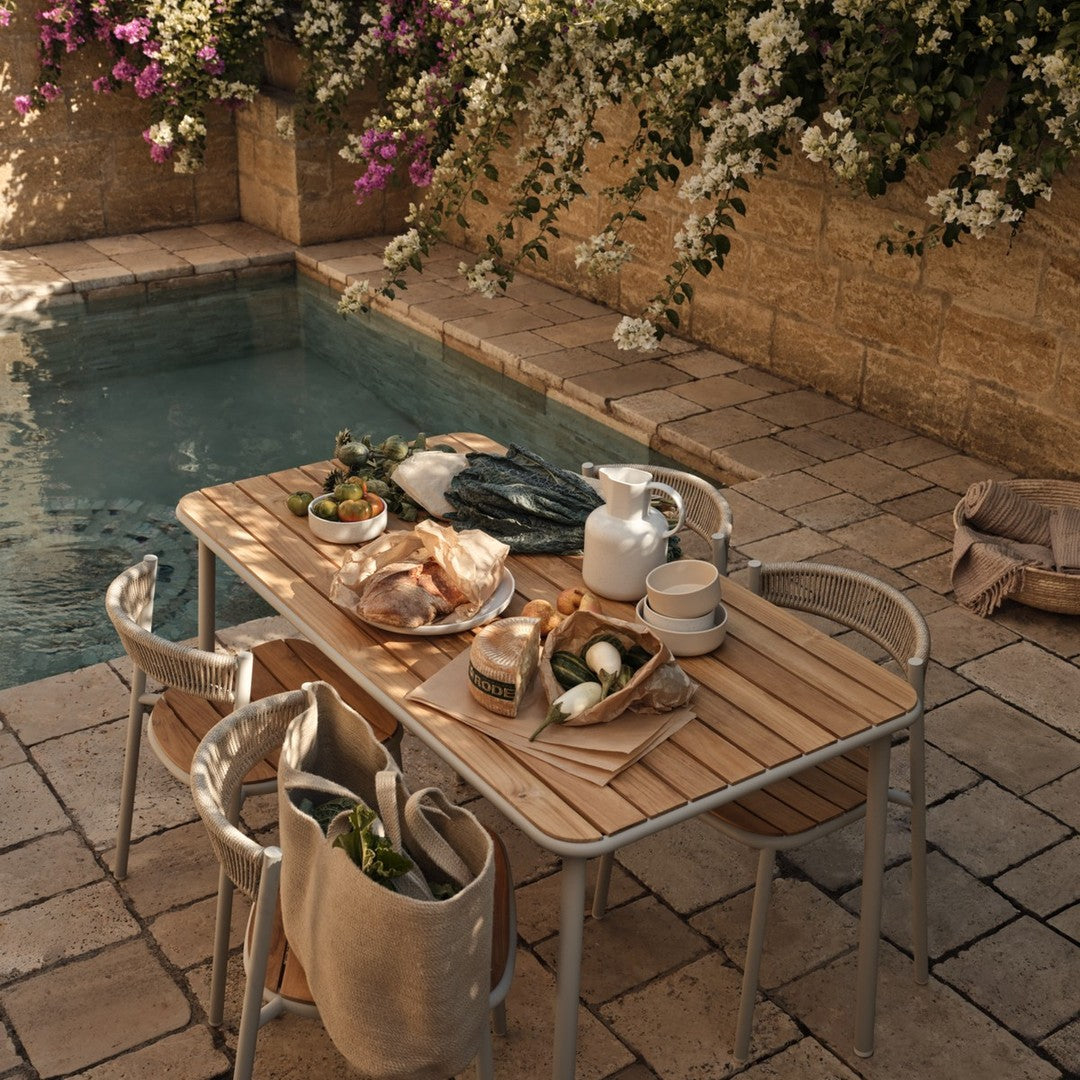Outdoor dining table with natural wood finish, neutral tones, and subtle textures, surrounded by blooming purple flowers.