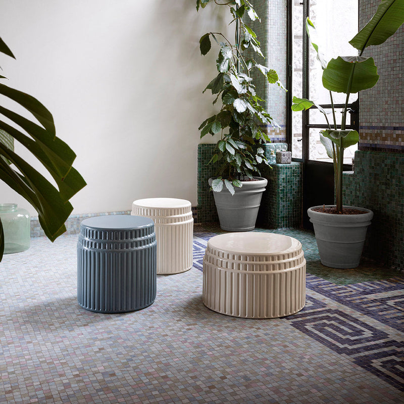 Three stylish ribbed stools in blue, white, and natural tones, set against patterned floor tiles and vibrant greenery.