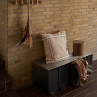 Neutral-toned striped pillow on a modern gray bench, paired with a warm brown circular stool. Soft, inviting textures.