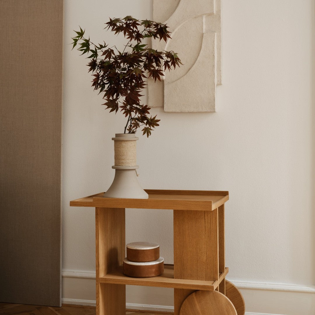 Natural wood cart with warm beige vase and red-leafed plant against soft muted wall, featuring minimalist design elements.