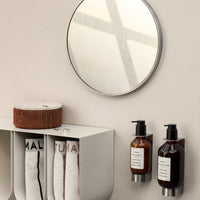 Round mirror reflecting light, black and amber bottles on a white shelf, and white towels with black lettering.
