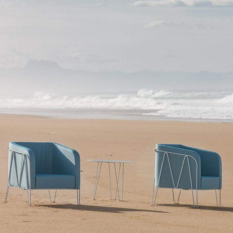 Two modern light blue chairs with sleek metal legs and a small white table, set against a beach background.