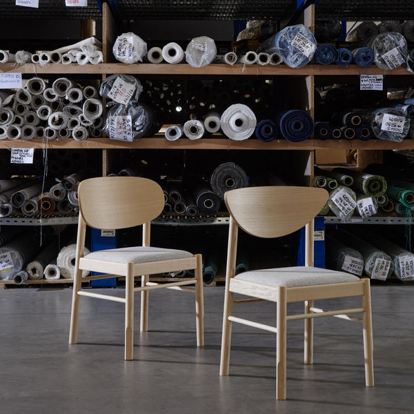 Set of two minimalist chairs featuring light wooden frames and white upholstered seats, against a backdrop of colorful fabric rolls.