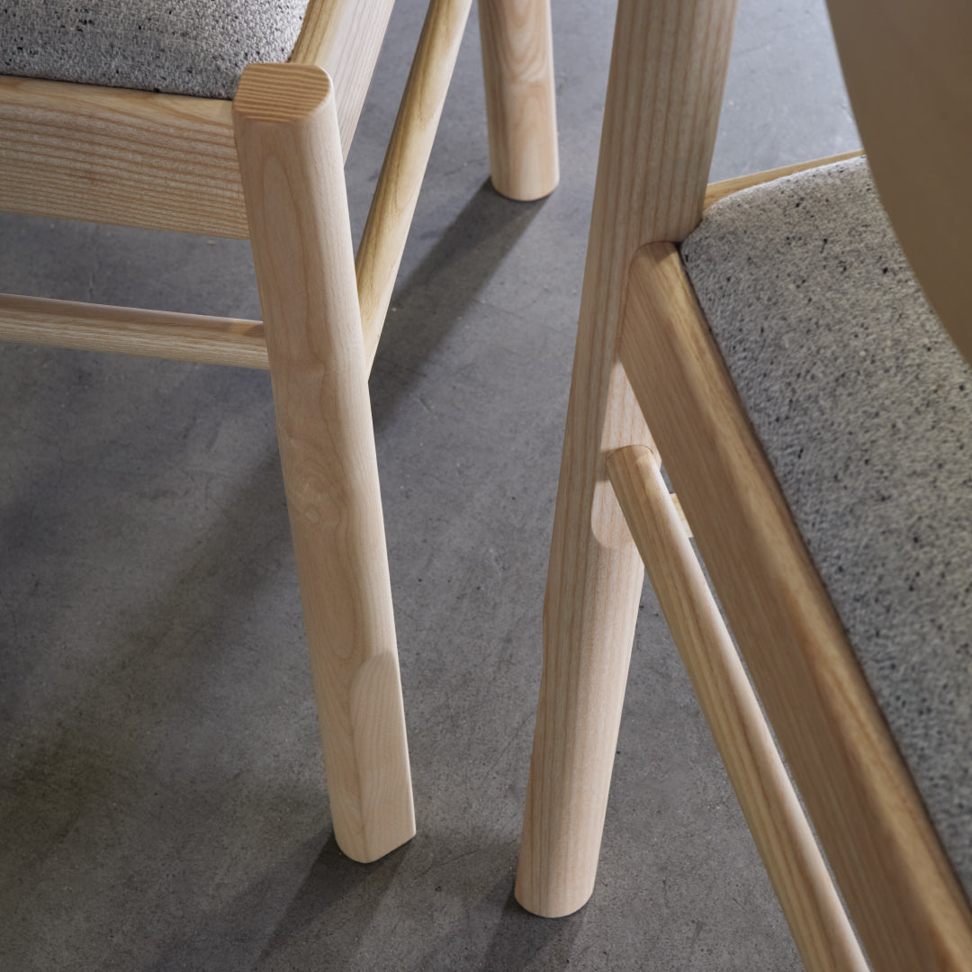 Light wood chair legs with a soft gray fabric seat against a sleek concrete floor. Clean, modern aesthetic.